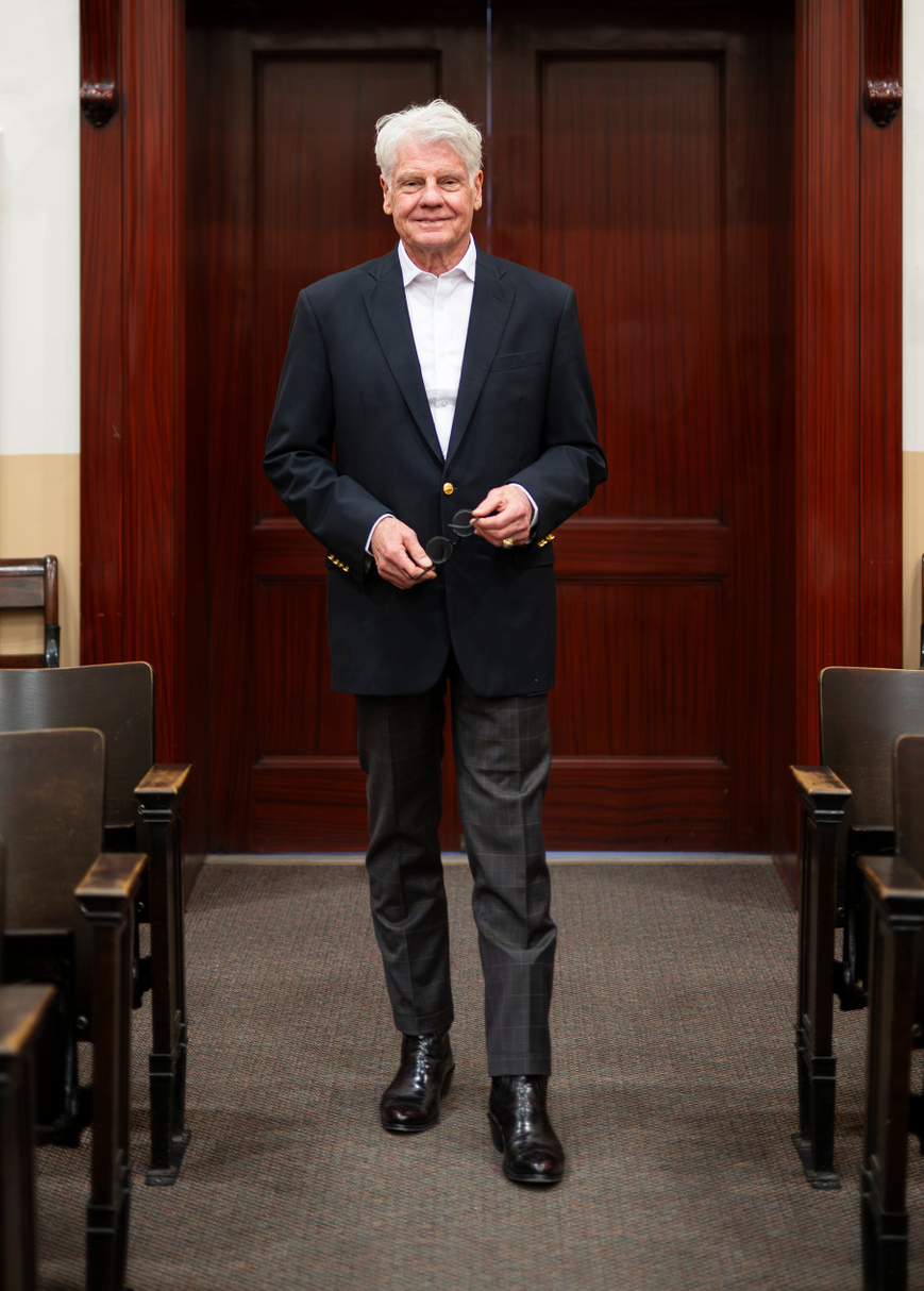 Distinguished older man standing in a courtroom, holding glasses with confidence.