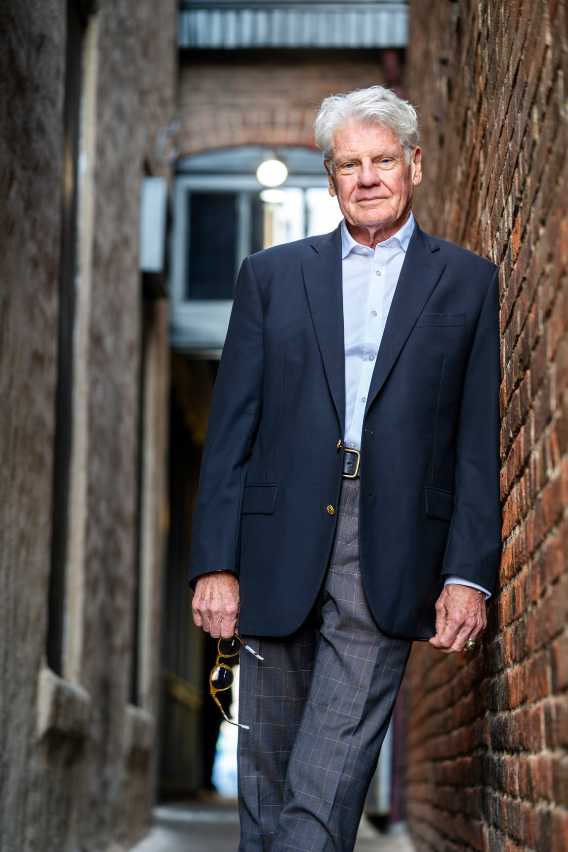 Distinguished older man in a suit leaning against a brick wall in a narrow alley.